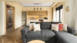Un salon moderne avec un canapé gris, un parquet en bois disposé en chevrons, une cuisine avec des meubles en bois et un éclairage suspendu.