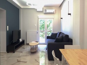 Een moderne woonkamer met een bank, een televisie, een tafeltje en een raam met uitzicht op groen.