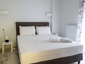 Una cama bien hecha con sábanas blancas, una mesita de noche con una lámpara y una ventana con cortinas florales.