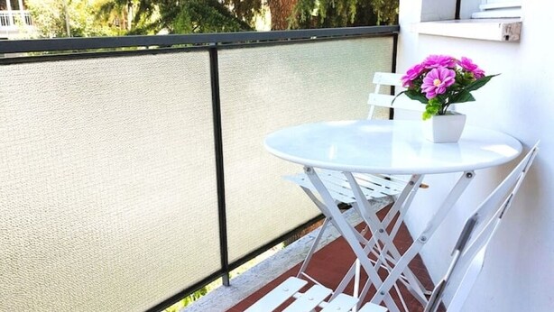 A small outdoor seating area with a round table and white chairs, a potted plant with pink flowers, and a view of trees through a textured fence. - Apartment Gioia: comfort and relaxation just minutes from Maria Cecilia Hospital (Lugo)