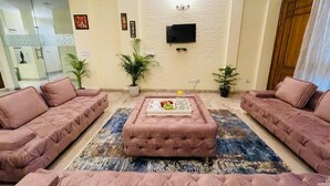 A living room with a large TV mounted on a brick wall, two sofas, a coffee table, and two potted plants. - Fortune Home Service Apartment 4Bhk D-36 (New Delhi)