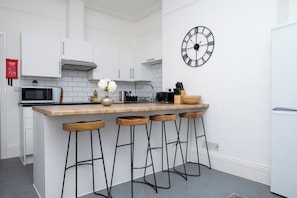 Una cocina moderna con armarios blancos, una isla de madera con taburetes de bar, un reloj negro y un microondas.