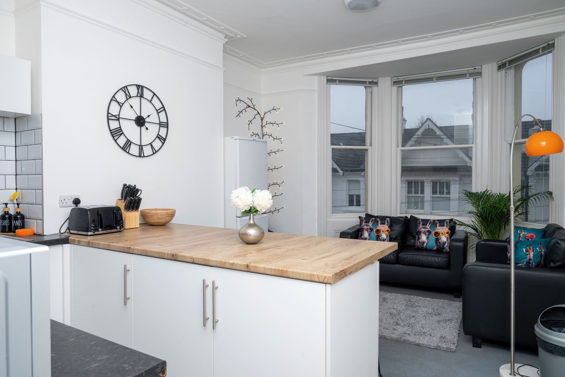 Una cocina moderna con encimera de madera, armarios blancos, un reloj negro y una zona de comedor con sofá negro y cojines decorativos.