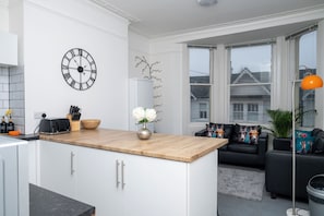 A modern kitchen with a wooden countertop, white cabinets, a black clock, and a dining area with a black sofa and decorative pillows.