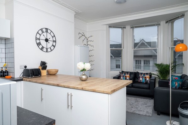 Una cocina moderna con encimera de madera, armarios blancos, un reloj negro y una zona de comedor con sofá negro y cojines decorativos.