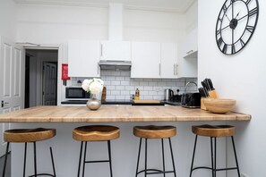 A modern kitchen with white cabinets, a wooden island with stools, a clock, and various kitchen appliances.