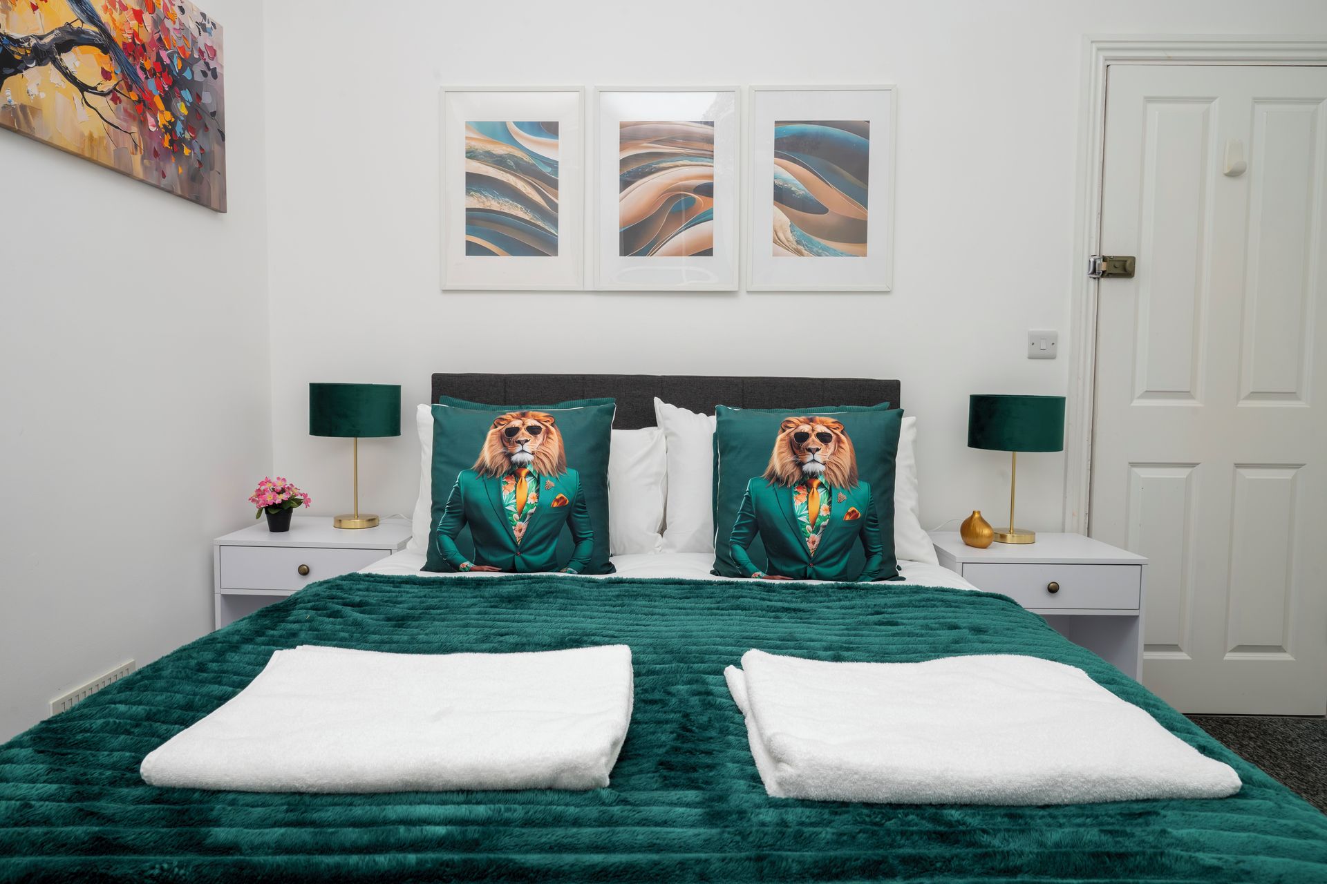 A bed with a green duvet, two pillows with a lion design, a nightstand with a lamp, and a wall with three framed artworks.