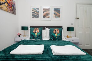 Una cama con edredón verde, dos almohadas con diseño de león, una mesita de noche con lámpara y una pared con tres obras de arte enmarcadas.