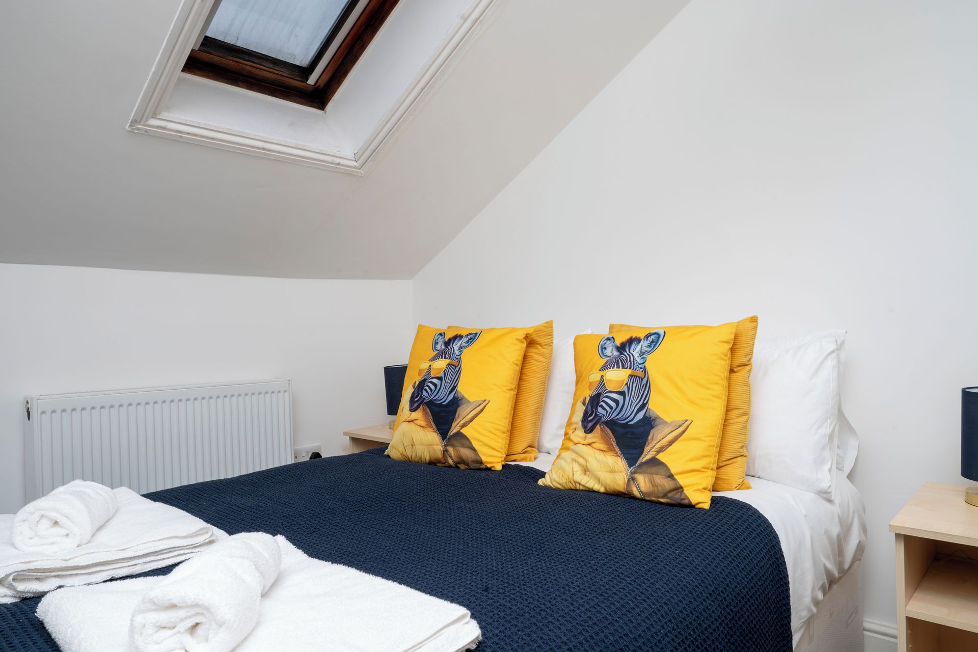 A bedroom with a skylight, a bed with dark blue bedding, two yellow pillows with zebra designs, a white radiator, and a wooden nightstand with a lamp.