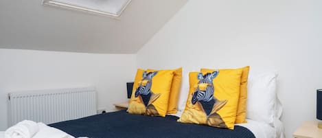 A bedroom with a skylight, a bed with dark blue bedding, two yellow pillows with zebra designs, a white radiator, and a wooden nightstand with a lamp.