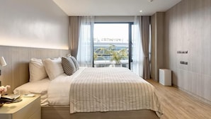 Una habitación de hotel moderna con una cama grande, una mesita de noche con lámpara, una planta pequeña en maceta y vistas a la ciudad a través de amplias ventanas.