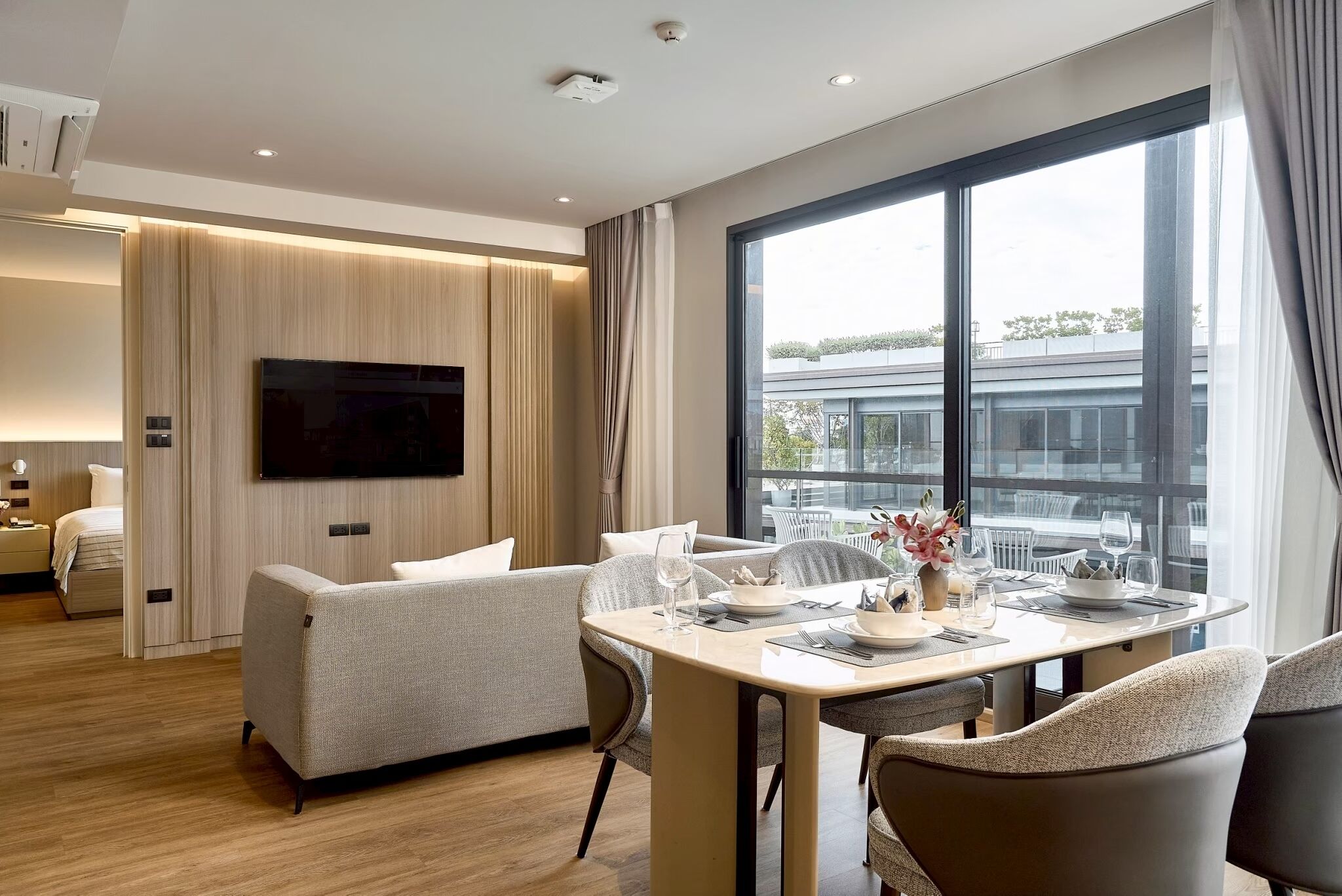 A modern hotel room with a dining area, a sofa, a flat-screen TV, and a large window offering a view of the city.