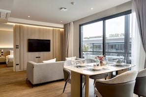 Une chambre d’hôtel moderne dotée d’un coin repas, d’un canapé, d’une télévision à écran plat et d’une grande fenêtre donnant sur la ville.