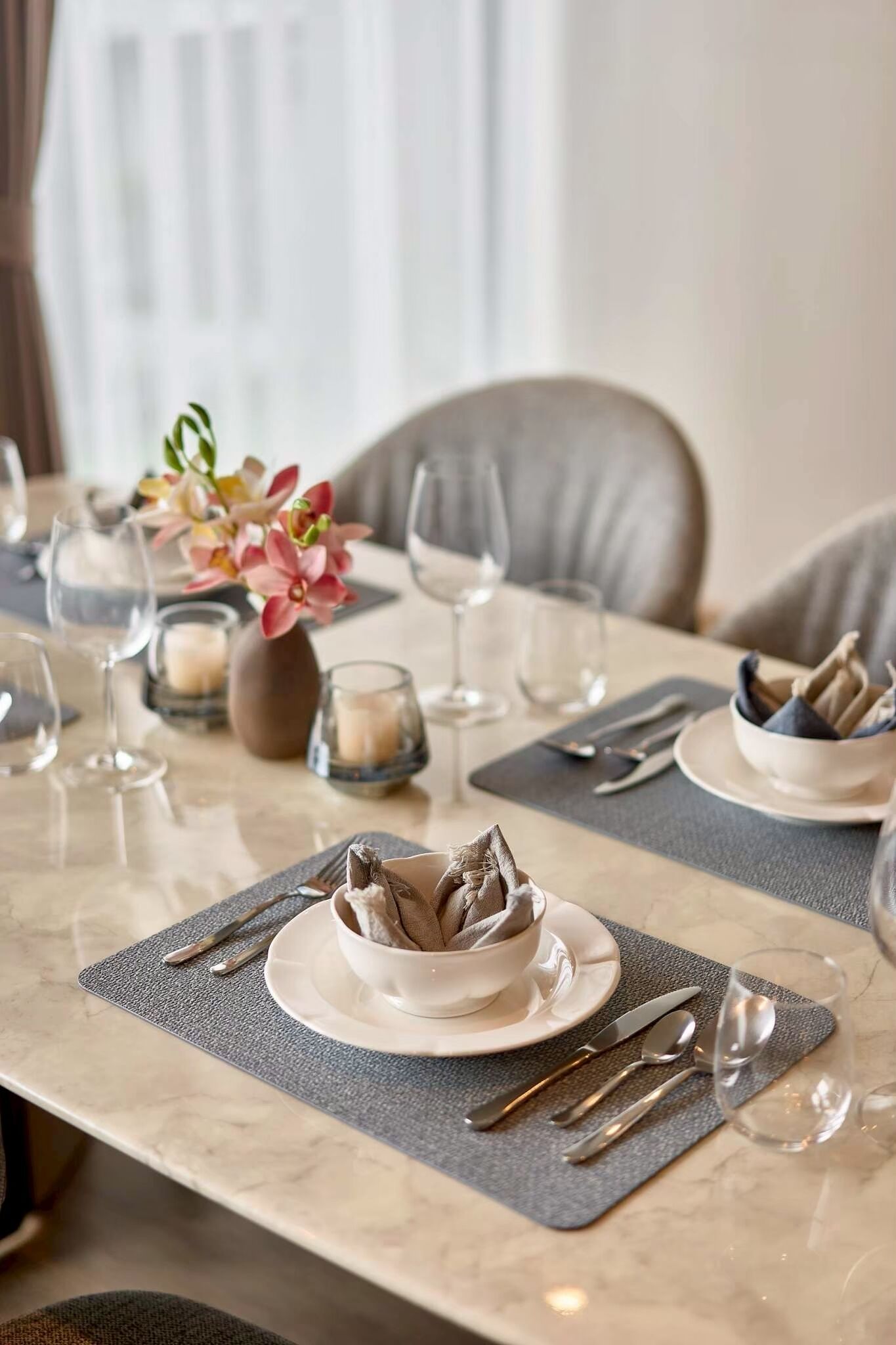 A dining table set with plates, cutlery, glasses, and candles, with a vase of flowers in the background.
