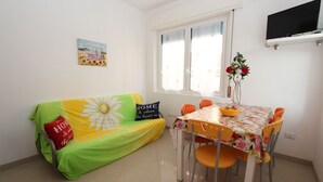 Una sala pequeña con sofá, una mesa de comedor con sillas, televisor montado en la pared y ventana con cortinas.