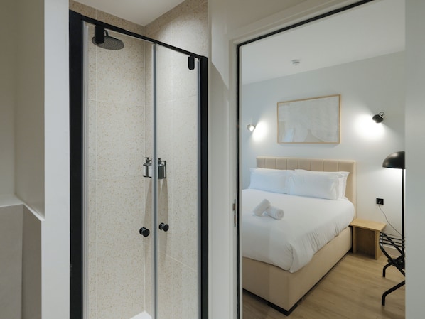 A modern hotel room with a glass shower enclosure, a bed with white linens, a bedside table, and a wall-mounted light fixture.