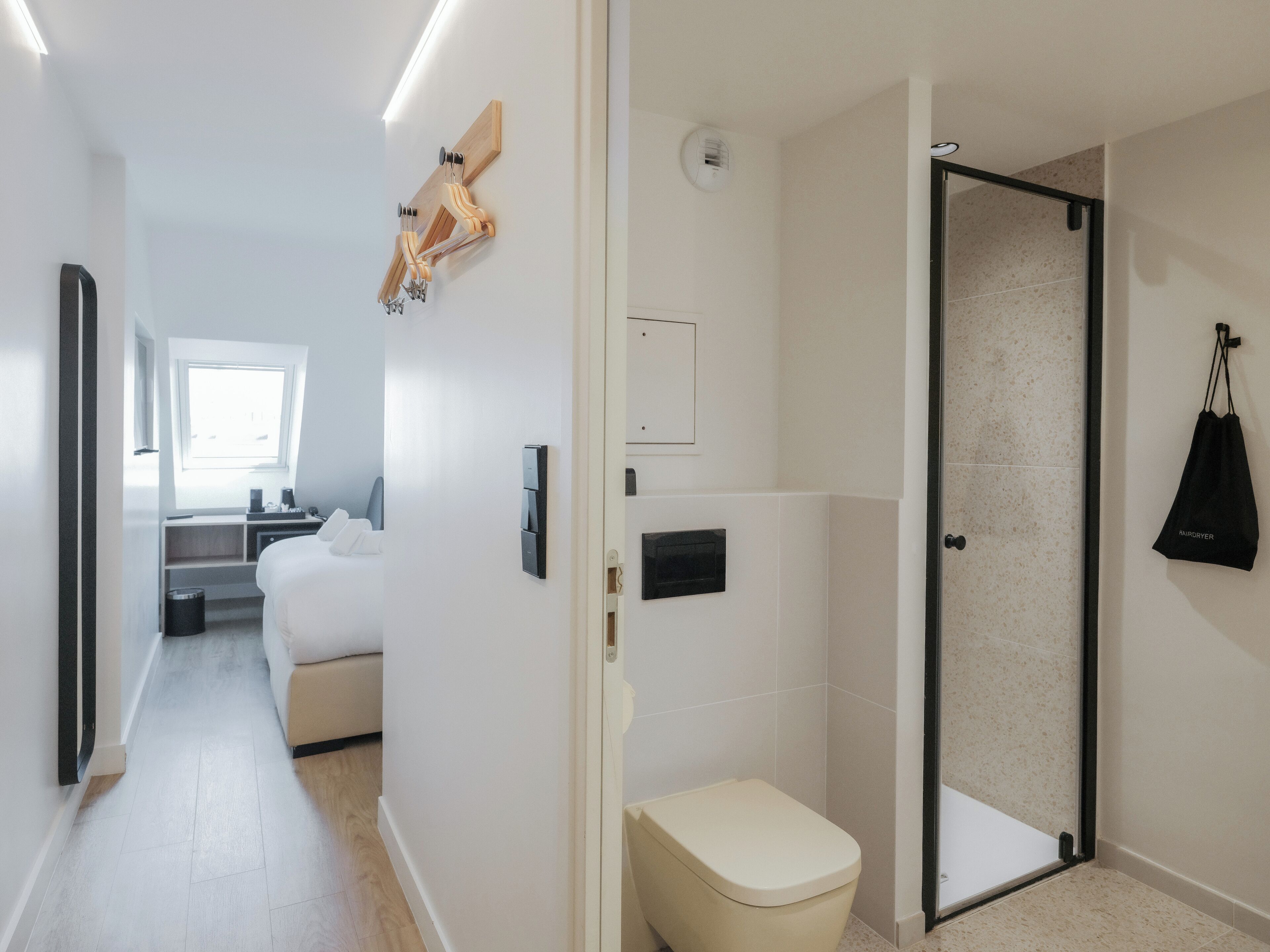 A modern hotel room with a bed, a bedside table, a window, a shower area, and a hanging bag.