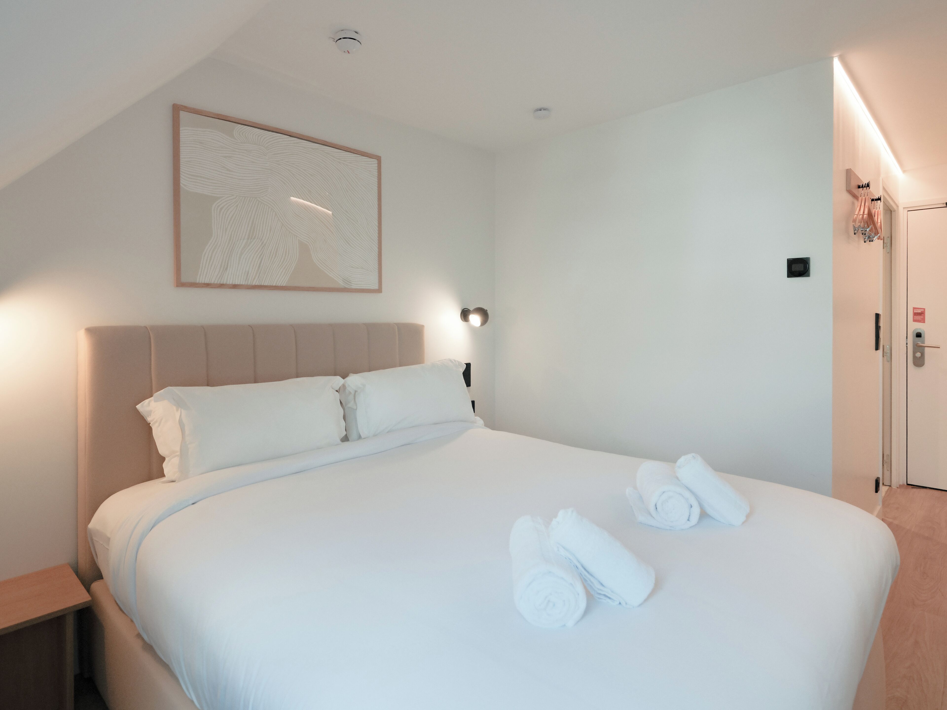 A hotel room with a large bed, a headboard, a bedside table, a wall-mounted light, a framed artwork, and a door with a towel rack.