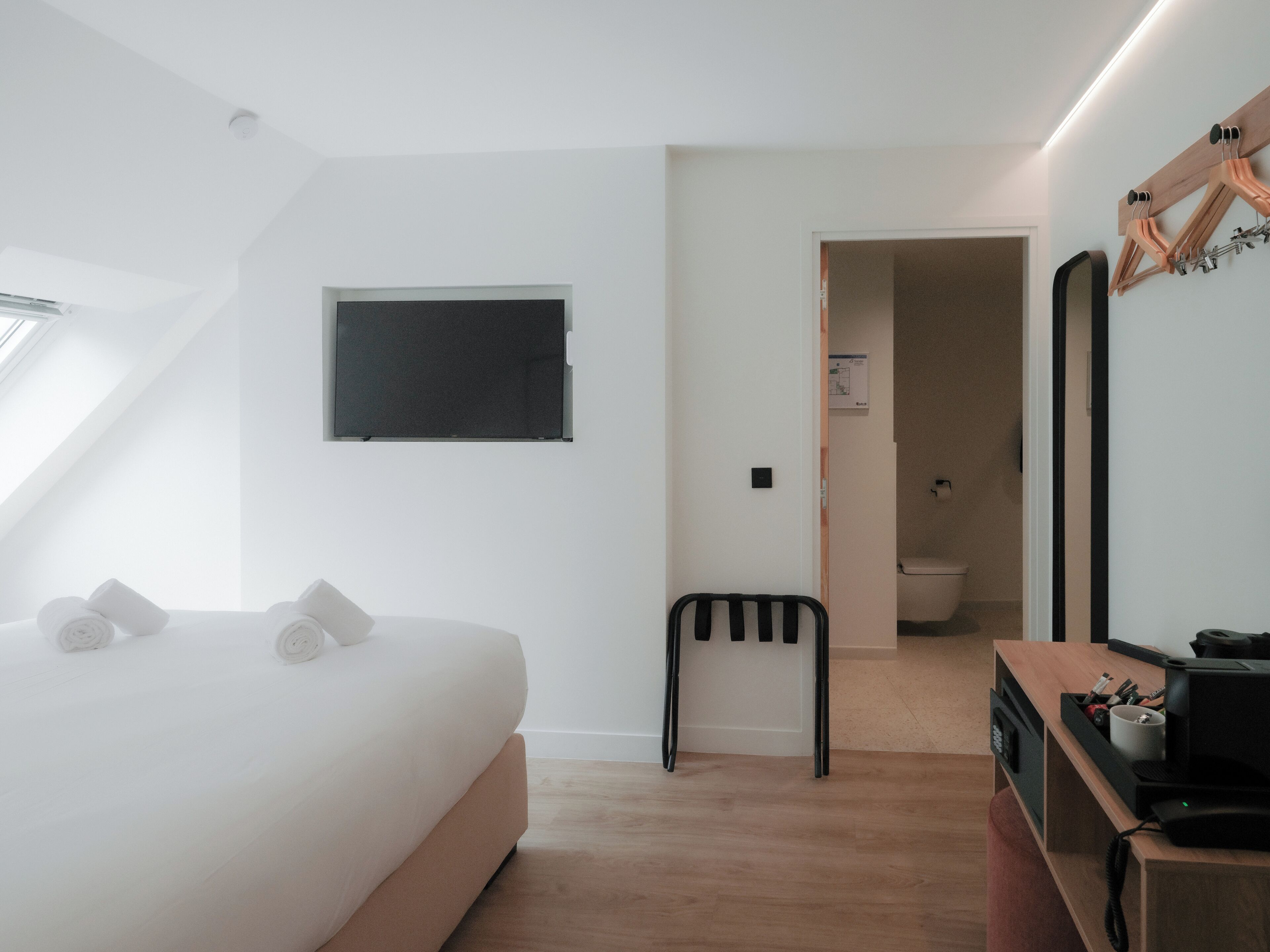 A modern hotel room with a bed, a desk, a chair, a small table, a mirror, and a window.