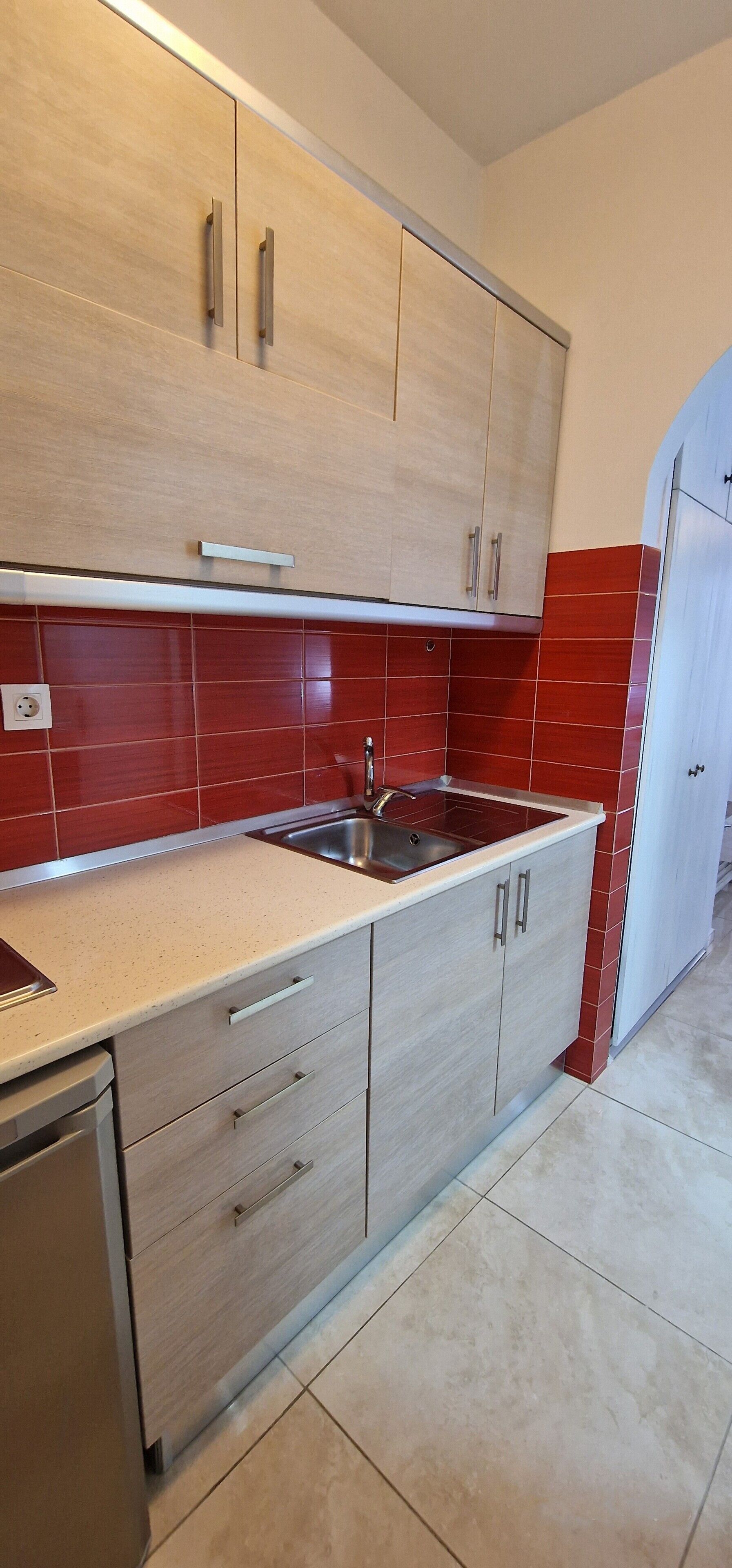 Una cocina con armarios de madera clara, un lavamanos de acero inoxidable y salpicadero con azulejos rojos.