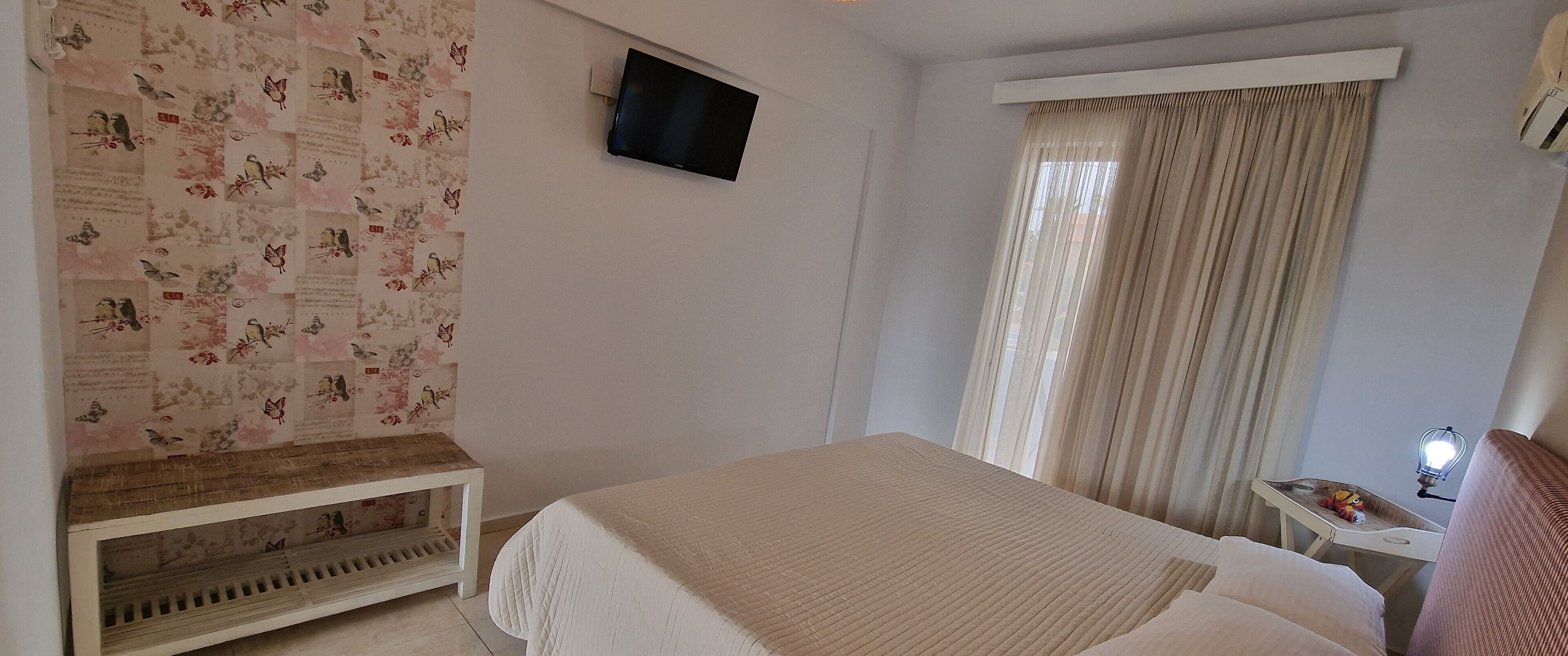 A bedroom with a bed, a TV mounted on the wall, a window with curtains, and a small table with a lamp.