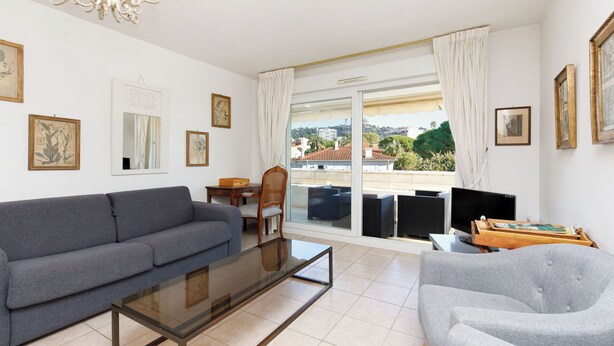 Ein modernes Wohnzimmer mit einem grauen Sofa, einem Glastisch und einem grauen Sessel. Es gibt eine Essecke mit einem Holztisch und Stühlen, und ein großes Fenster mit Blick auf die Stadt.