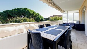 Eine überdachte Terrasse im Freien mit Tisch und Stühlen, Blick auf Bäume und ein Gebäude, sowie ein Deckenventilator.