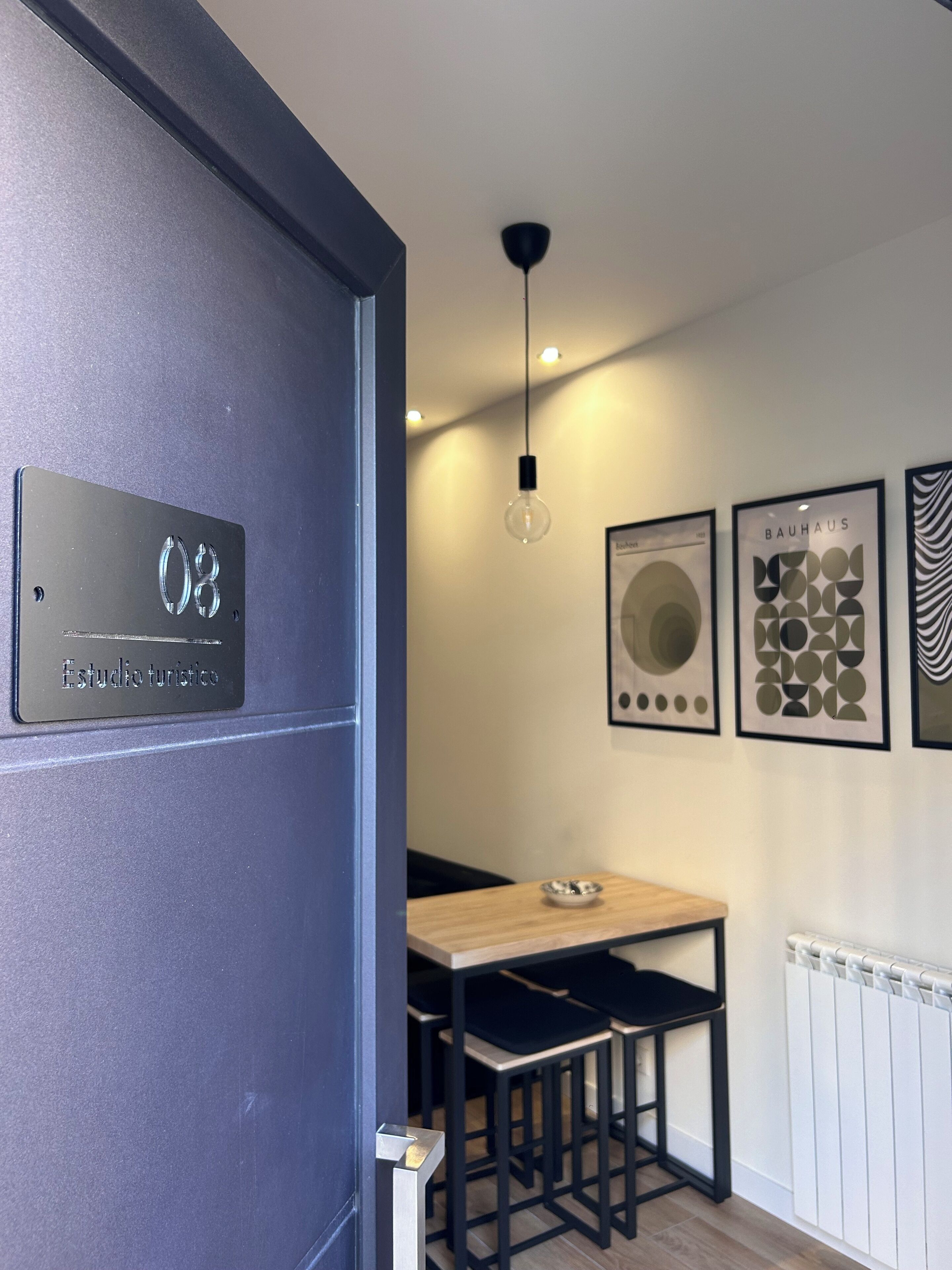 Un interior moderno con una puerta identificada como '08 Estudio Turistico', una zona de comedor con una mesa de madera y sillas negras, y tres obras de arte enmarcadas en la pared.