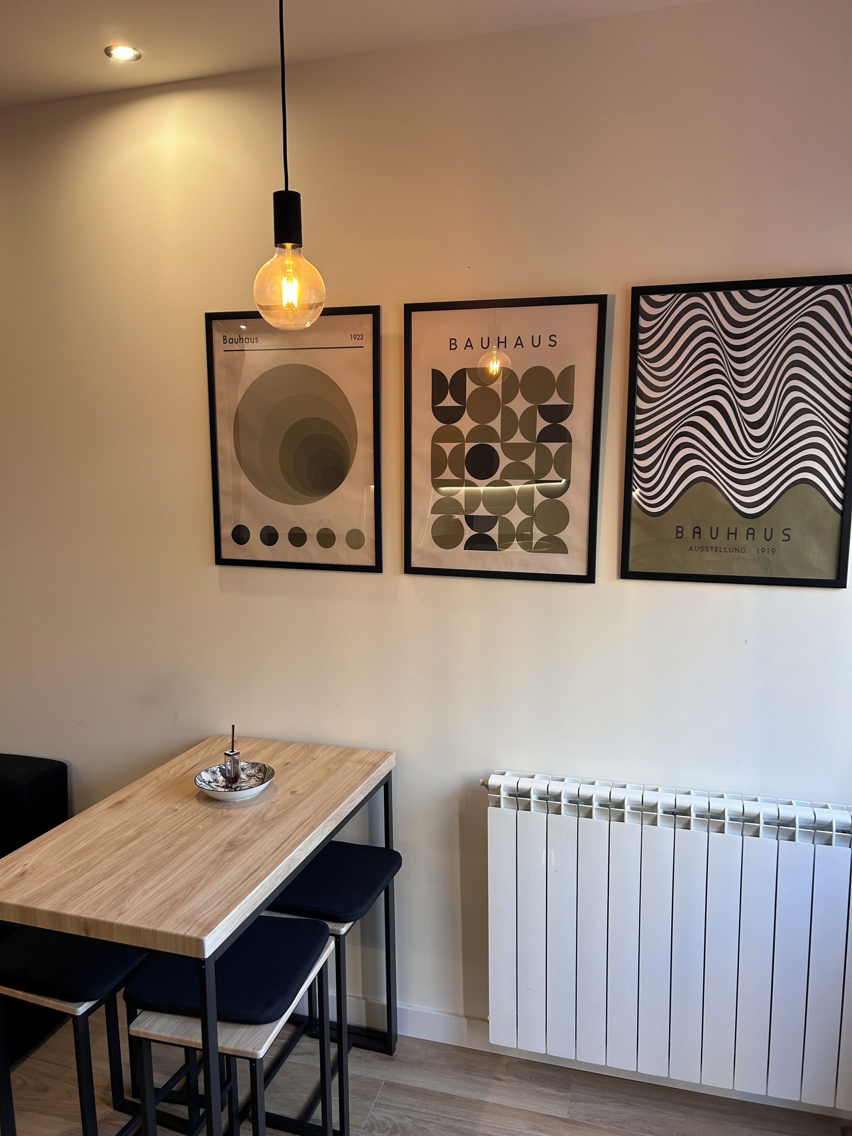 Un comedor con una mesa de madera, sillas negras, un lámpara colgante y tres obras de arte Bauhaus enmarcadas.