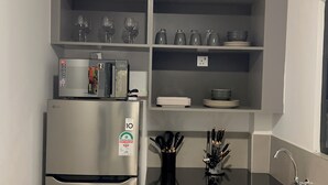 Une cuisine équipée d’un réfrigérateur en acier inoxydable, d’un four à micro-ondes et d’une machine à café posés sur le plan de travail. Des étagères situées au-dessus servent à ranger des verres à vin, des assiettes et des bols.