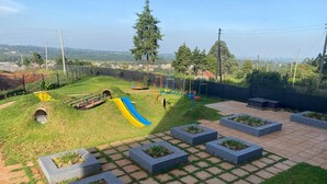 Un terrain de jeux équipé de toboggans et de balançoires, entouré d’une pelouse agrémentée de jardinières et offrant une vue sur des bâtiments éloignés.