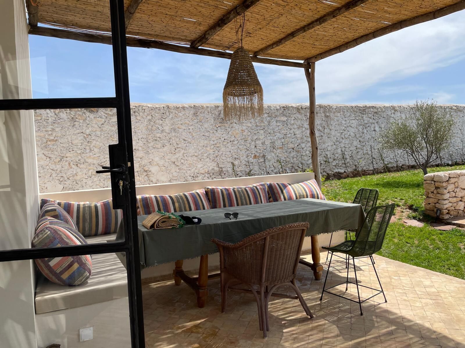 Un área de estar al aire libre con una mesa, sillas, techo tejido y pared de piedra al fondo.