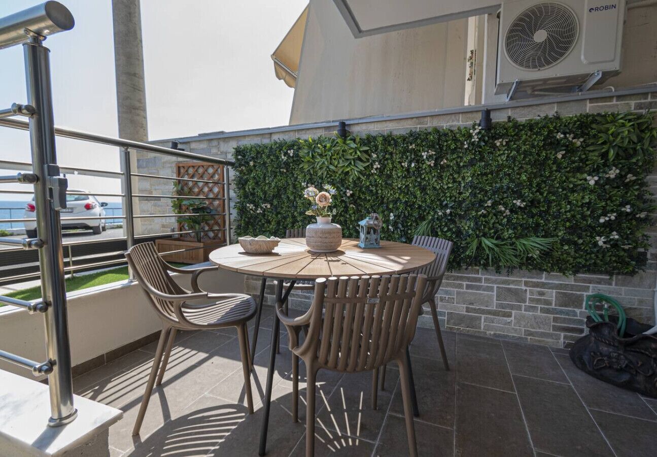 A round table with chairs, a decorative vase, and a lantern on a balcony with a green wall and a view of the sea.