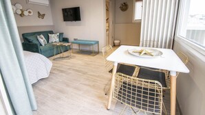 Ein Zimmer mit Bett, Fernseher, Esstisch mit Stühlen und einem Fenster mit Jalousien.