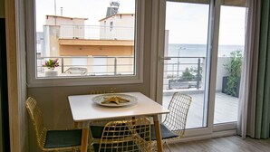Ein Zimmer mit Tisch, zwei Stühlen, einer Meeressterne als Dekoration, einem Fenster mit Blick auf Gebäude und das Meer sowie einem Balkon mit Geländer.
