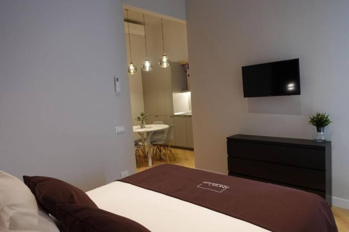 A bedroom with a bed, a dresser, a TV mounted on the wall, and a dining area in the background.