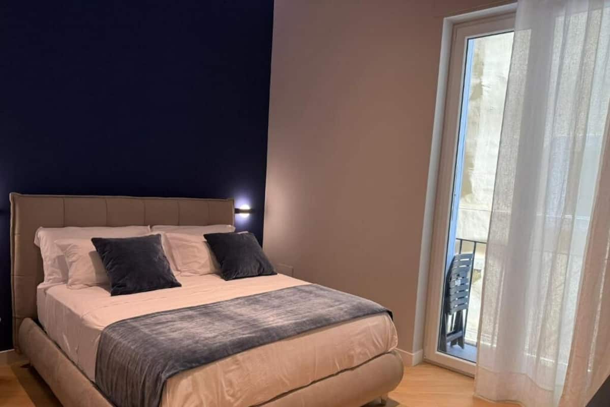 A bedroom with a bed, a nightstand, a window with curtains, and a view of the outdoors.