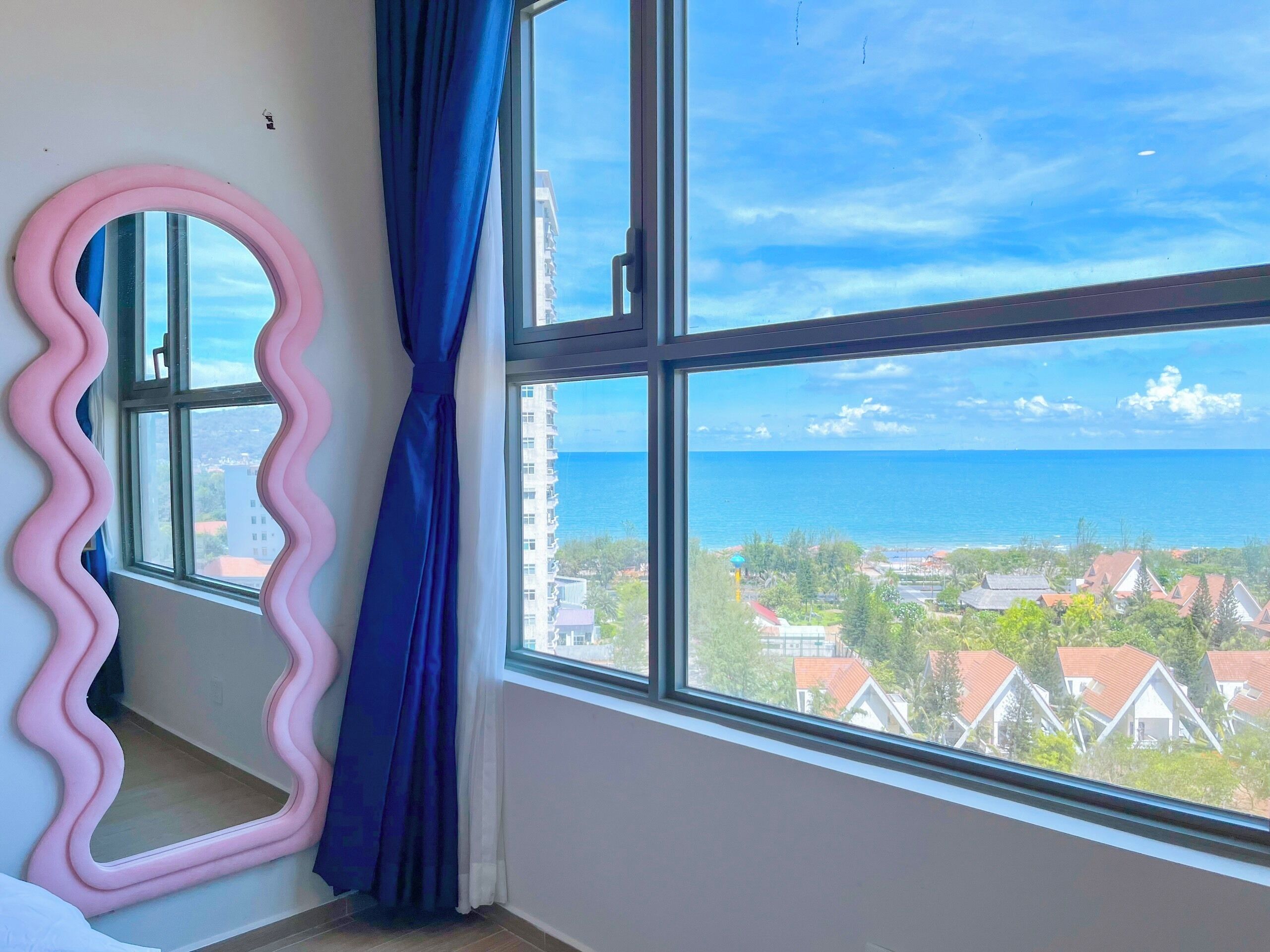 Una habitación con un ventanal grande, un espejo ondulado rosa y una cortina azul.