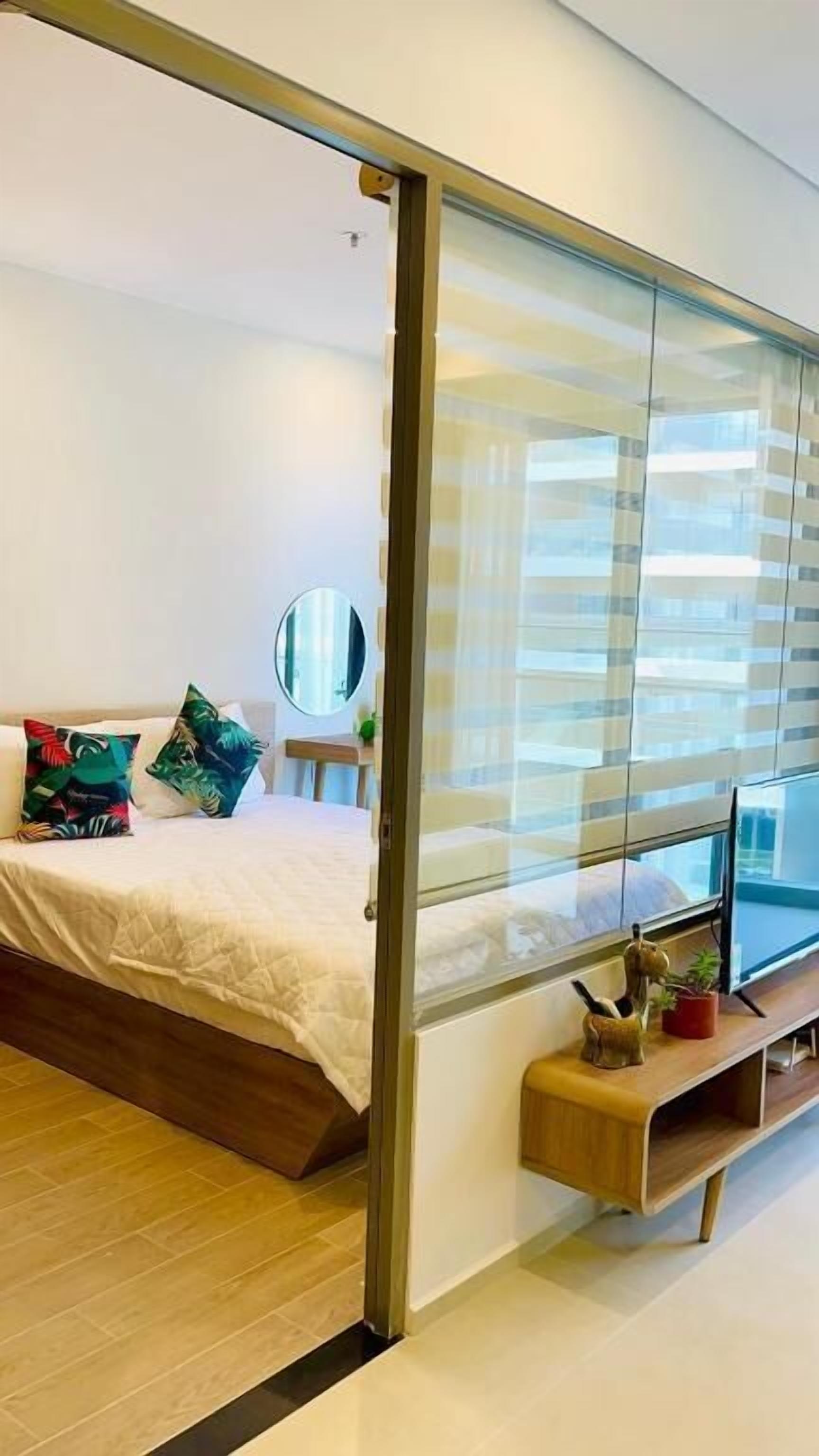 A modern bedroom with a bed, a bedside table with a mirror, a TV, and a sliding glass door leading to a balcony.