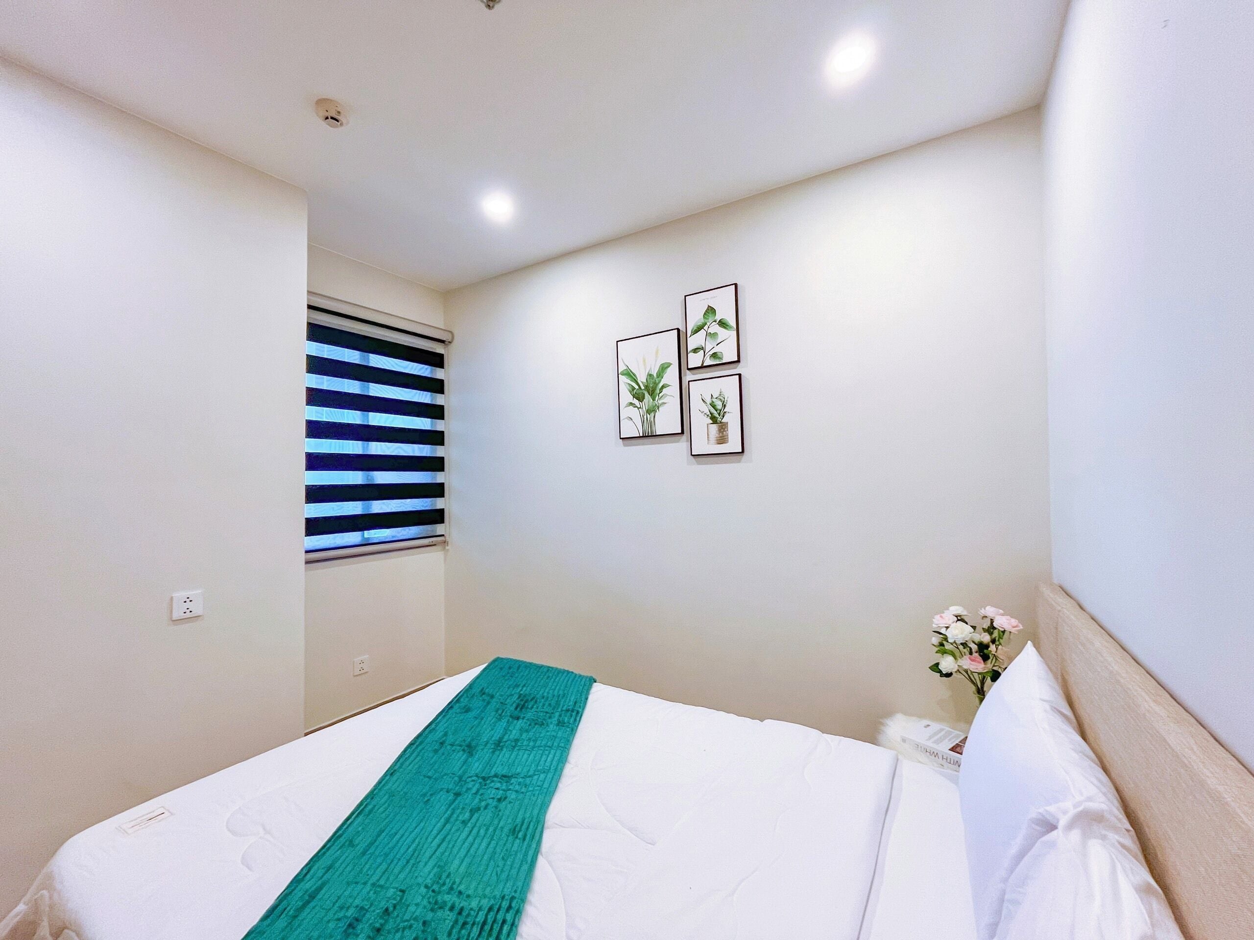 A bedroom with a bed, a window with blinds, a wall-mounted light, and two framed pictures.