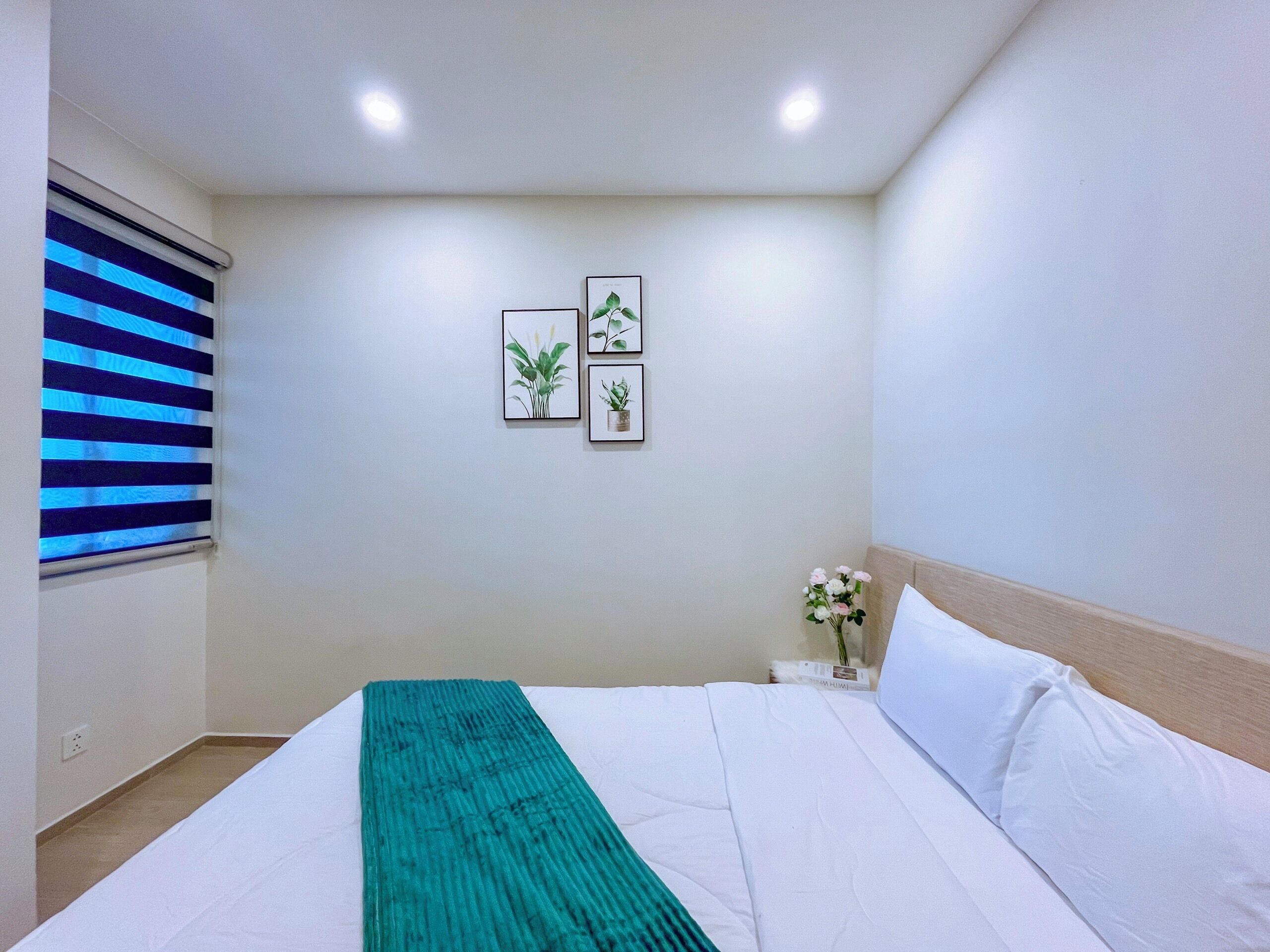A bedroom with a bed, a nightstand, a window with blinds, and two framed botanical prints on the wall.