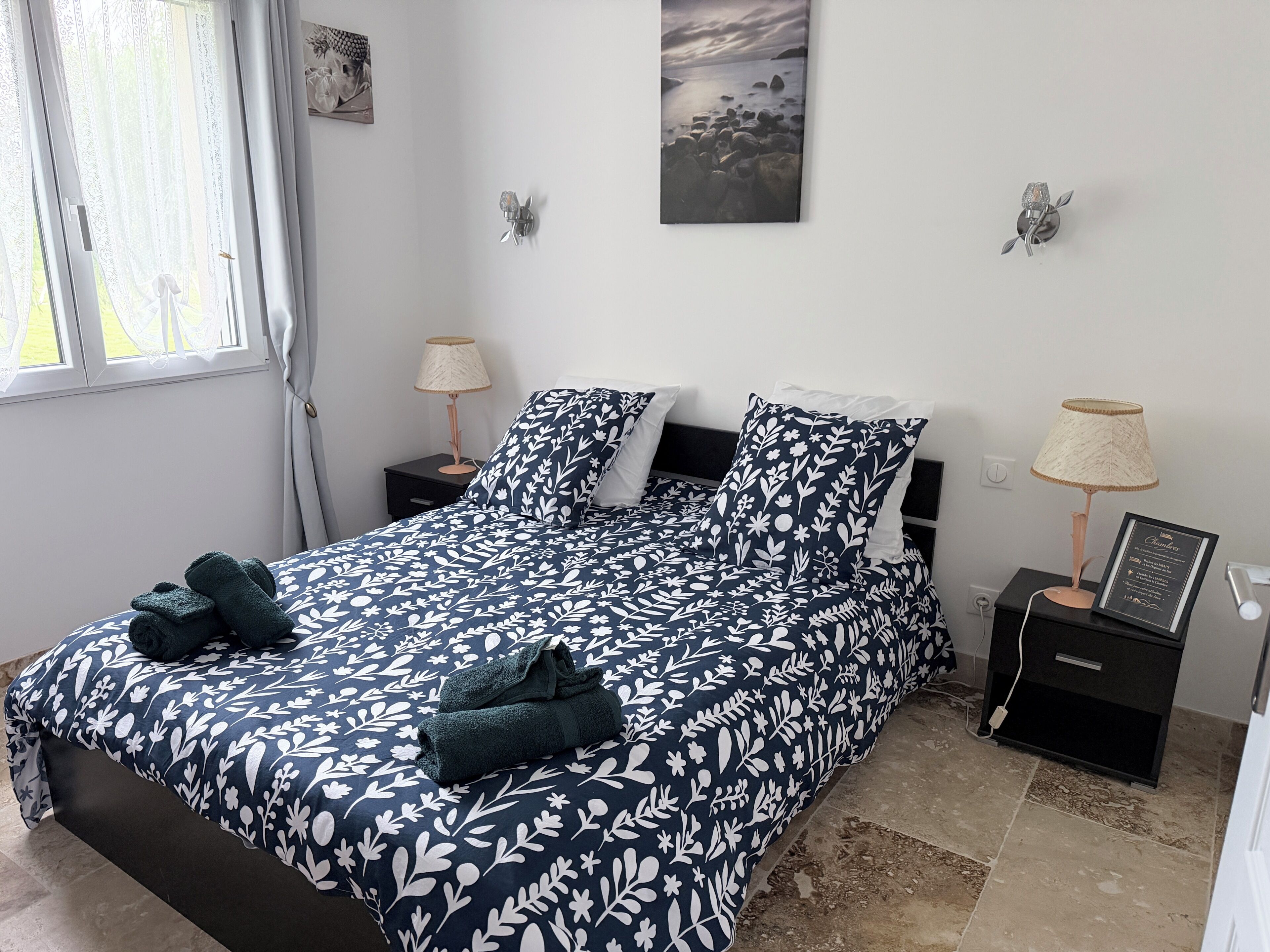 Una cama bien hecha con una colcha floral, una lámpara de noche y un cuadro en la pared.