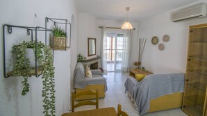 Ein kleiner, gemütlicher Raum mit einer Couch, einem Sessel, einem Bett und einem kleinen Tisch. Es gibt ein Fenster mit Vorhängen, eine hängende Pflanze und einen Spiegel an der Wand.