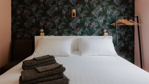 Un lit bien fait, avec du linge de lit blanc, une pile de serviettes pliées et un papier peint orné de motifs tropicaux.
