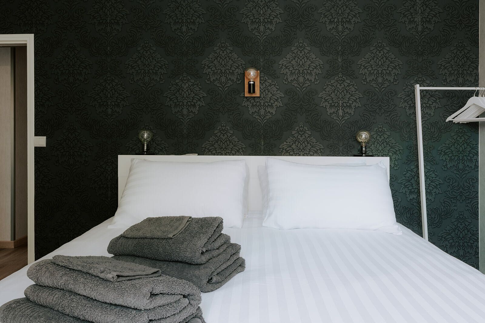A neatly made bed with white linens and folded towels, a wall with a floral pattern, and a small wall-mounted light fixture.