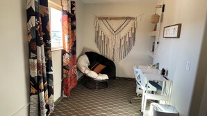 A compact room with a patterned curtain, a macrame wall hanging, a white chair, a small desk, and a trash bin. - Awesome 1-bedroom apartment with AC in charming Page (Page)