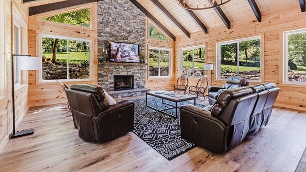 A cozy cabin interior with a stone fireplace, wooden walls, and large windows offering a view of the outdoors. - 6BR 63 Acre Park Like Estate Loud Creek Trails Pond Huge Yard Pickleball Golf (Whittier)