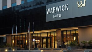 Un HOTEL WARWICK con una fachada moderna, una entrada de vidrio grande y un logo de corona prominente.