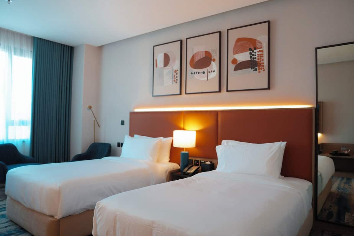 A hotel room with two beds, a nightstand with a lamp, a desk with a chair, and a large window with curtains.