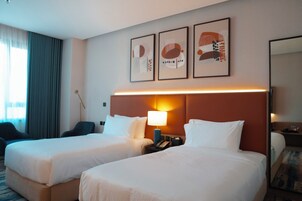 Habitación de hotel con dos camas, una mesita de noche con lámpara, un escritorio con silla y un ventanal con cortinas.